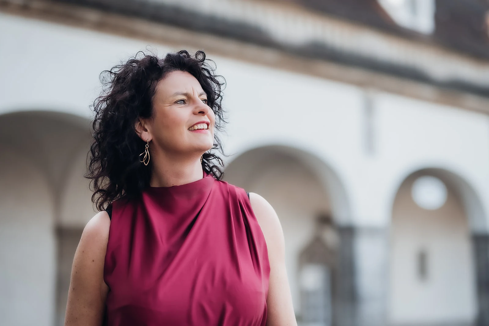 Martina Koch-Lang steht lächelnd im Freien vor historischen Arkaden und blickt zur Seite.